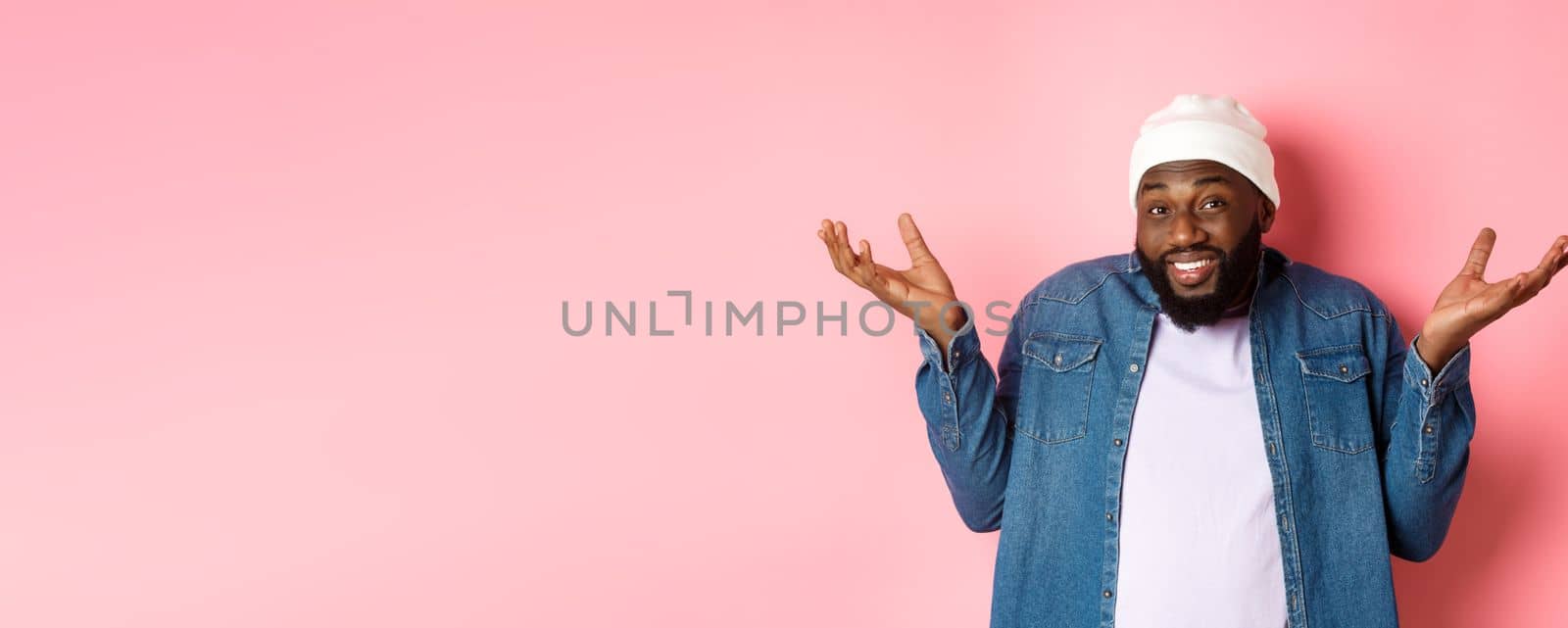 Unsure Black guy shrugging and looking confused at camera, dont know, standing indecisive against pink background.