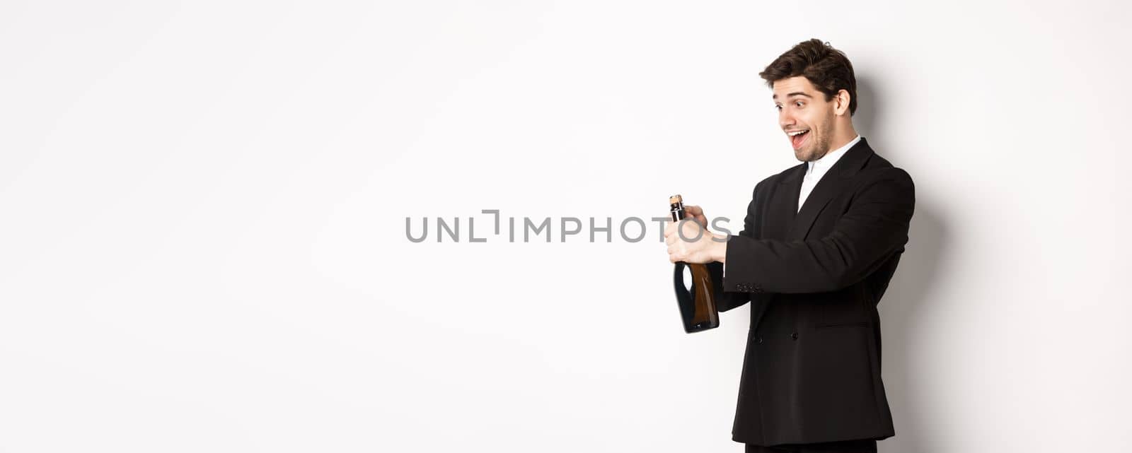 Portrait of attractive man in black suit, winking at camera and opening bottle of champagne, celebrating new year, standing against white background by Benzoix