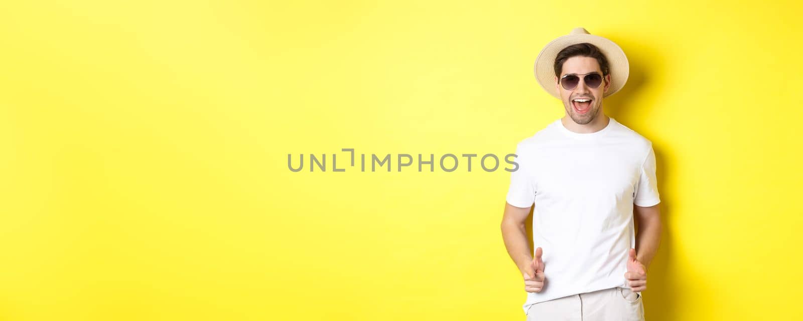 Confident and cheeky guy on vacation flirting with you, pointing finger at camera and winking, wearing summer hat with sunglasses, yellow background.