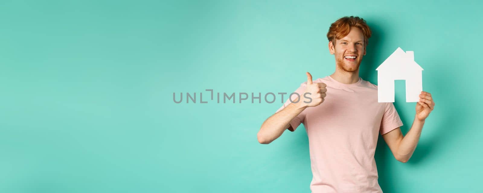 Real estate concept. Young man with red hair, wearing t-shirt, showing paper house cutout and thumb up, recommend agency, standing over turquoise background.
