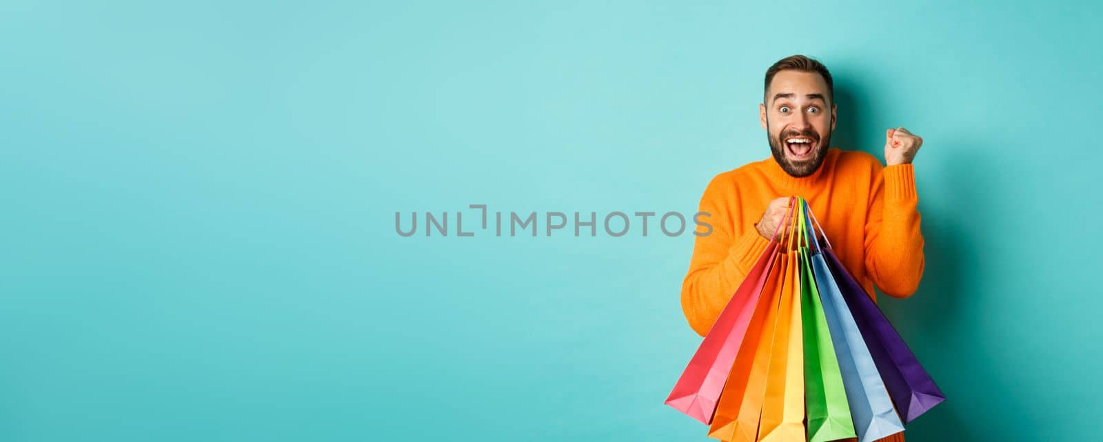 Lifestyle concept. Excited man showing shopping bags and rejoicing from discounts, standing over turquoise background.