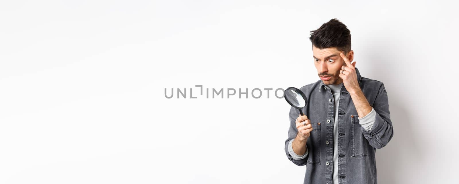 Man looking through magnifying glass and stretching eyelid to see clearly, standing on white background by Benzoix