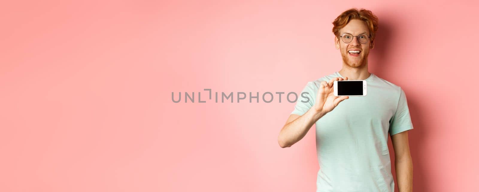 Happy young man with red hair showing smartphone screen, holding phone horizontally and smiling amazed, standing over pink background.