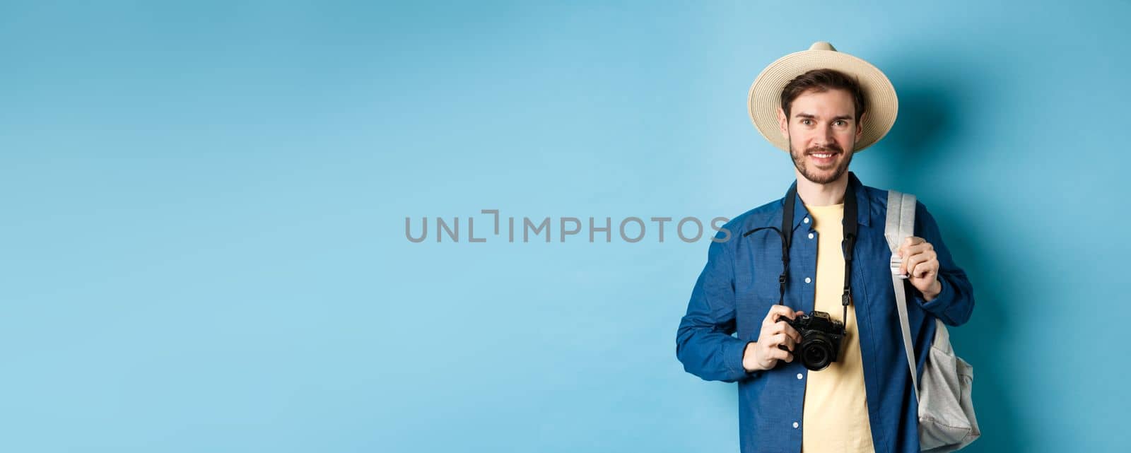 Cheerful handsome guy going on vacation, wearing summer hat and holding backpack with camera for photos, smiling excited of holiday, standing on blue background by Benzoix