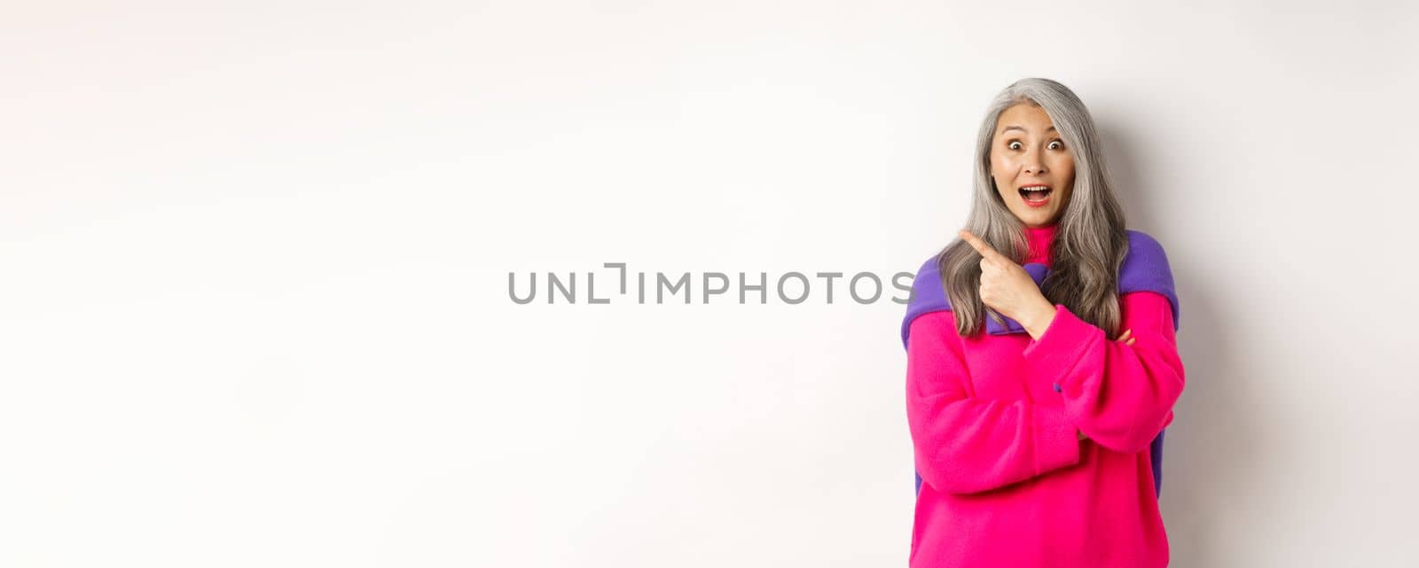 Portrait of impressed asian lady checking out promo offer, pointing at upper left corner and staring at camera amazed, standing in trendy pink sweater over white background.