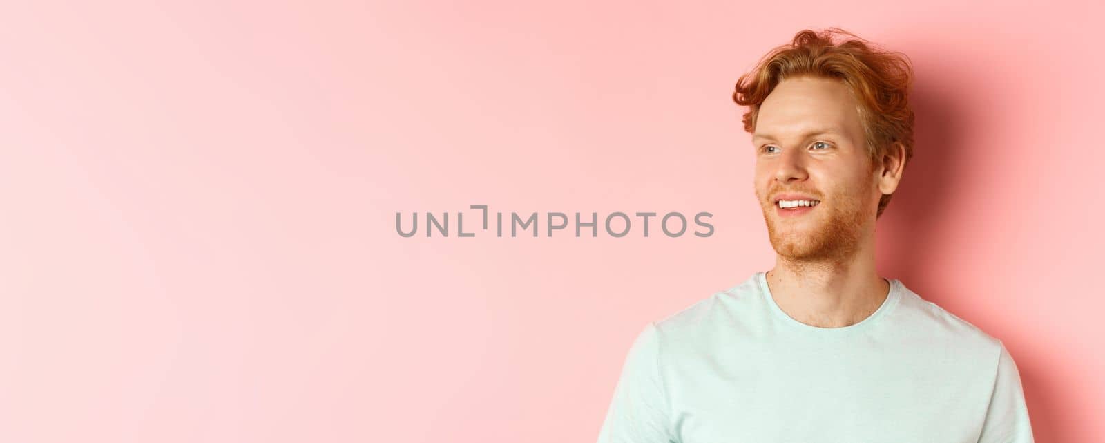 Portrait of attractive caucasian guy with red messy hair and beard, turn head and looking left with pleased smile, standing over pink background.