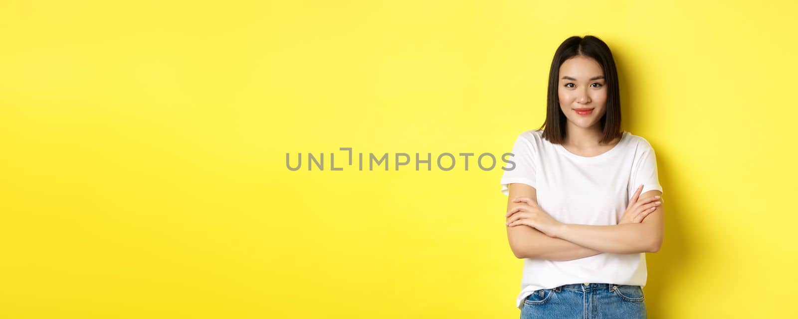 Confident and stylish asian woman cross arms on chest and smiling, standing over yellow background by Benzoix