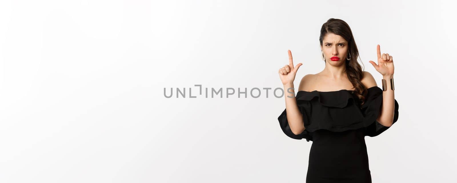 Fashion and beauty. Disappointed woman sulking upset, pointing fingers up and complaining, standing dissatisfied in black dress, white background.