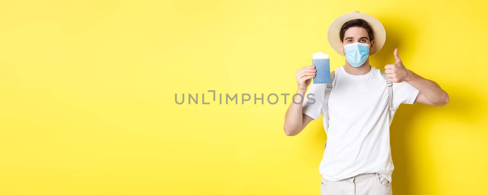 Concept of covid-19, tourism and pandemic. Happy male tourist in medical mask showing passport, going on vacation during coronavirus, make thumb up sign, yellow background by Benzoix