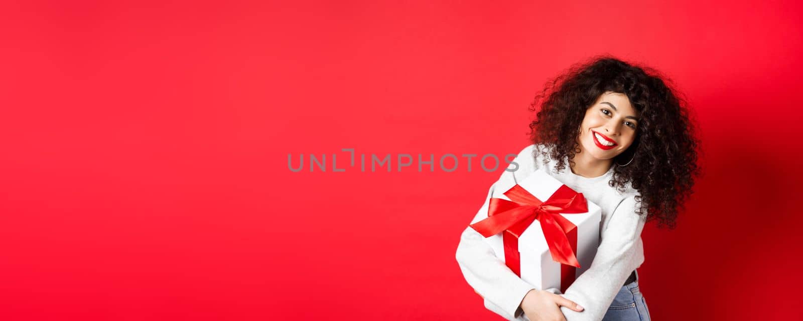 Celebration and holidays concept. Happy woman holding birthday gift and smiling at camera, standing in casual clothes, red background.