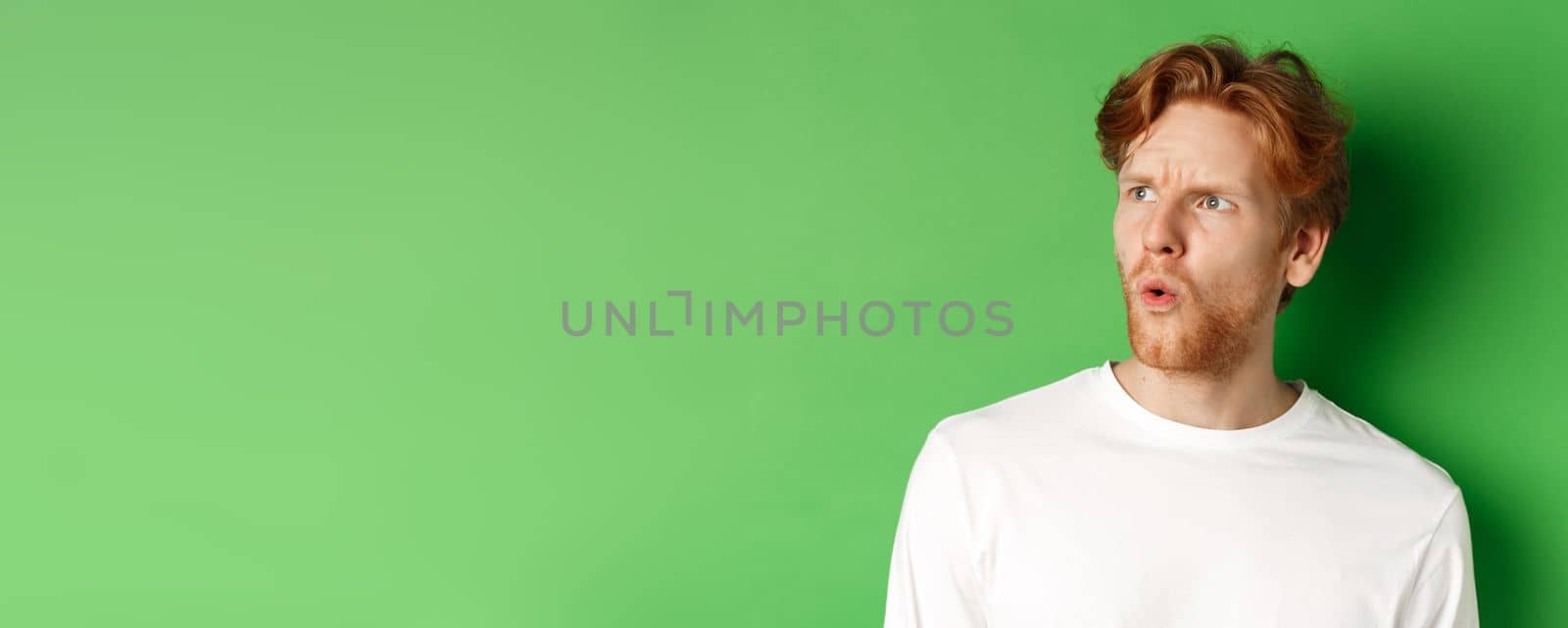 Confused redhead man staring left and looking worried, standing against green background.