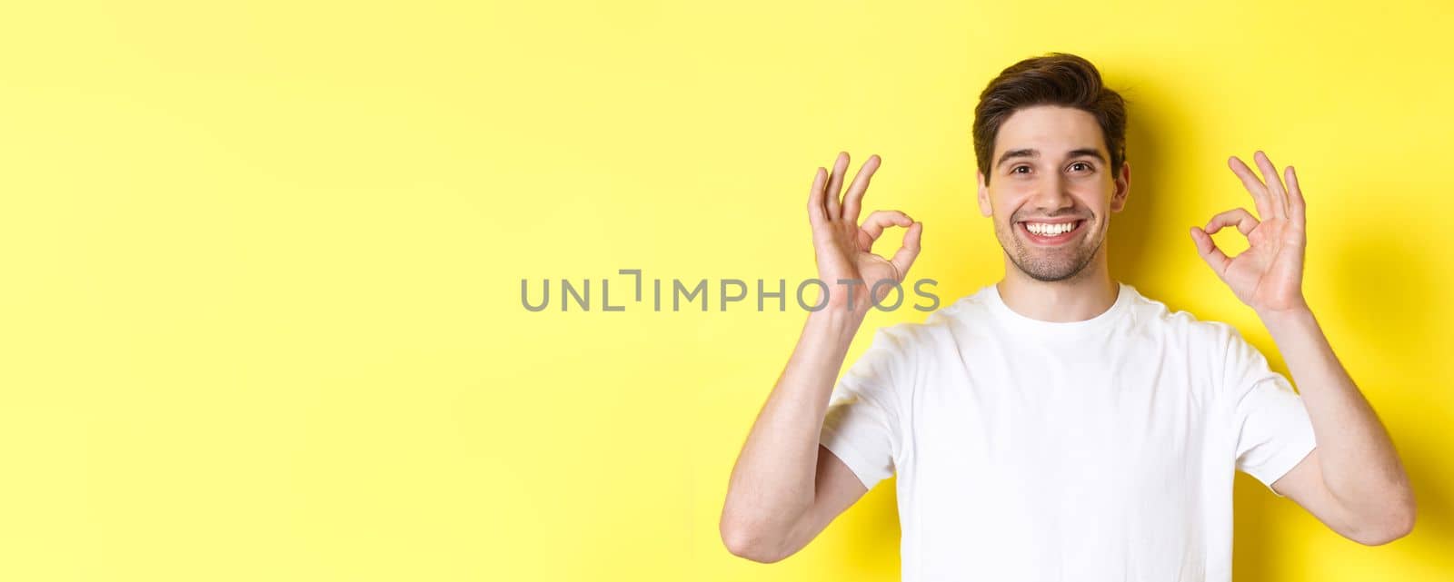 Close-up of handsome young man showing okay sign, approve and agree, smiling satisfied, standing over yellow background by Benzoix