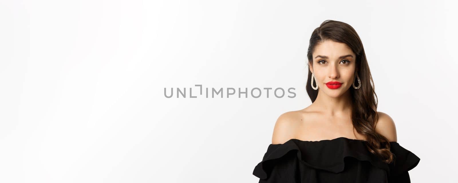 Fashion and beauty concept. Close-up of elegant brunette woman with earrings, wearing black dress and red lipstick, looking sensual at camera, standing over white background.