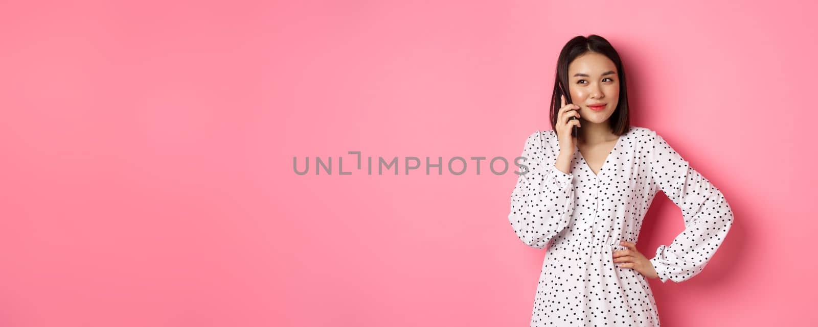 Attractive korean woman making a phone call, holding smartphone near ear and smiling, standing over pink background.