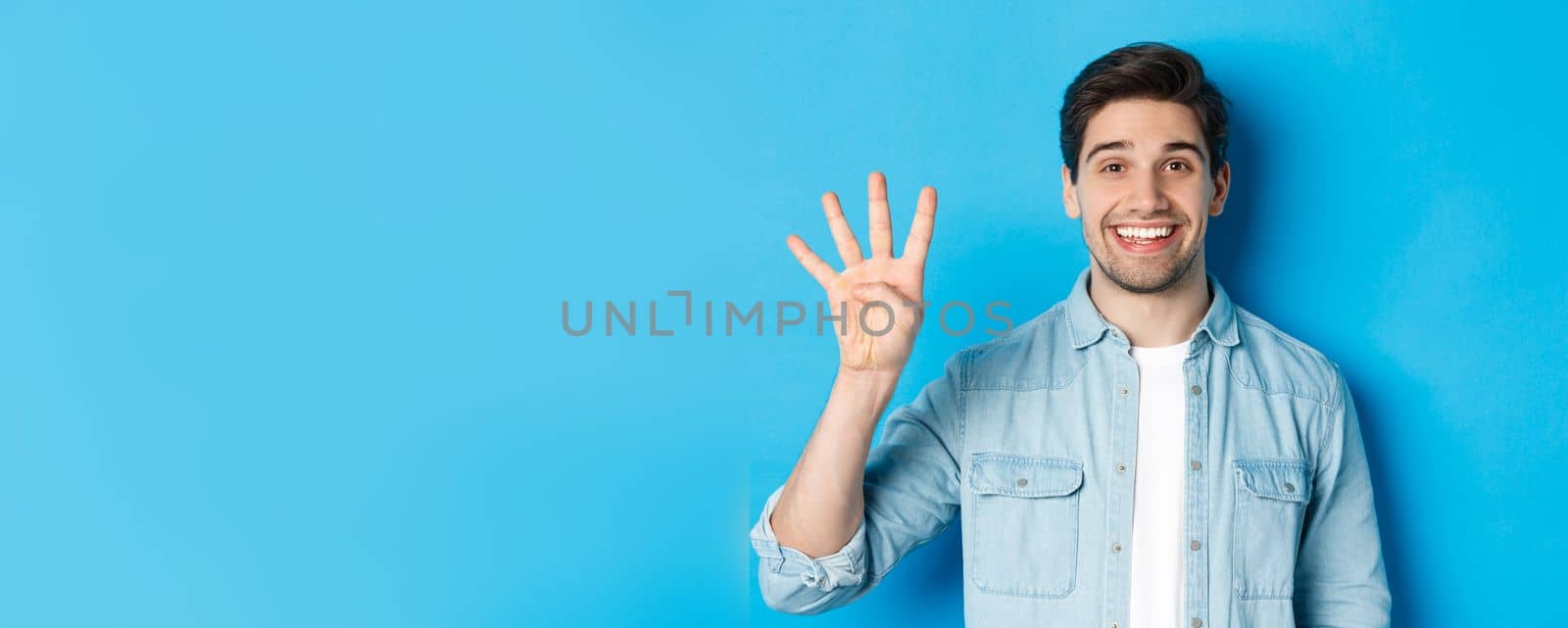 Close-up of handsome man smiling, showing fingers number four, standing over blue background by Benzoix