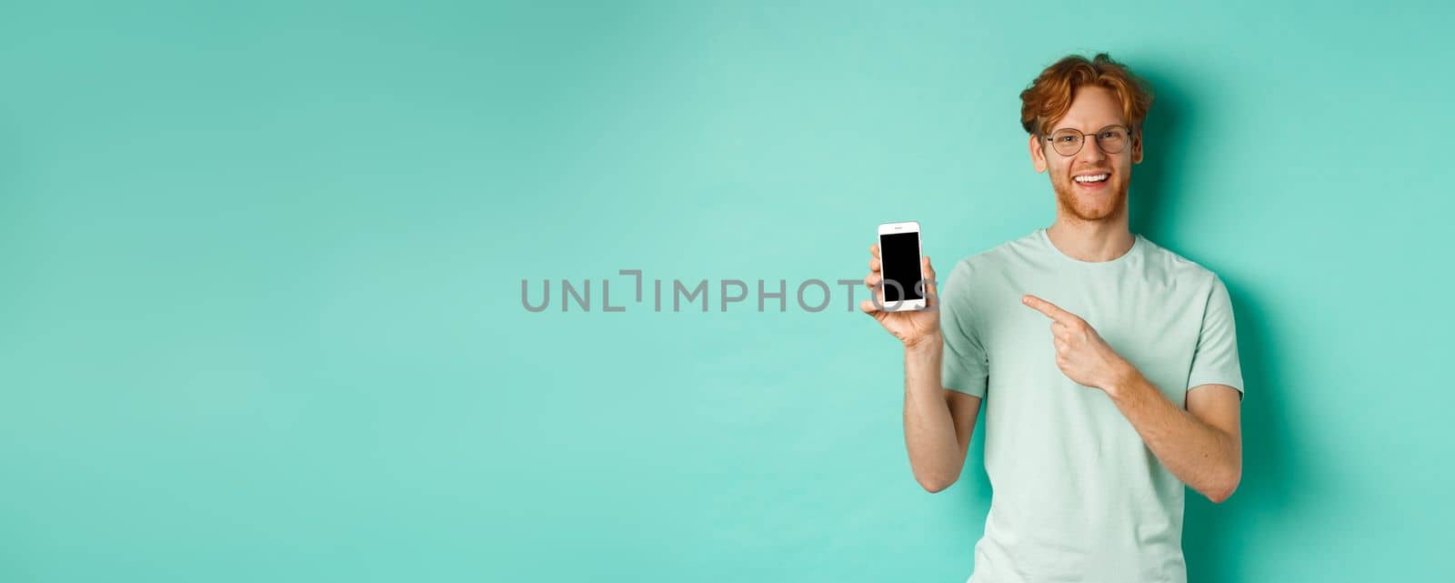 Attractive young man with red beard and hair pointing finger at blank smartphone screen, showing online promotion or app, smiling at camera, turquoise background.