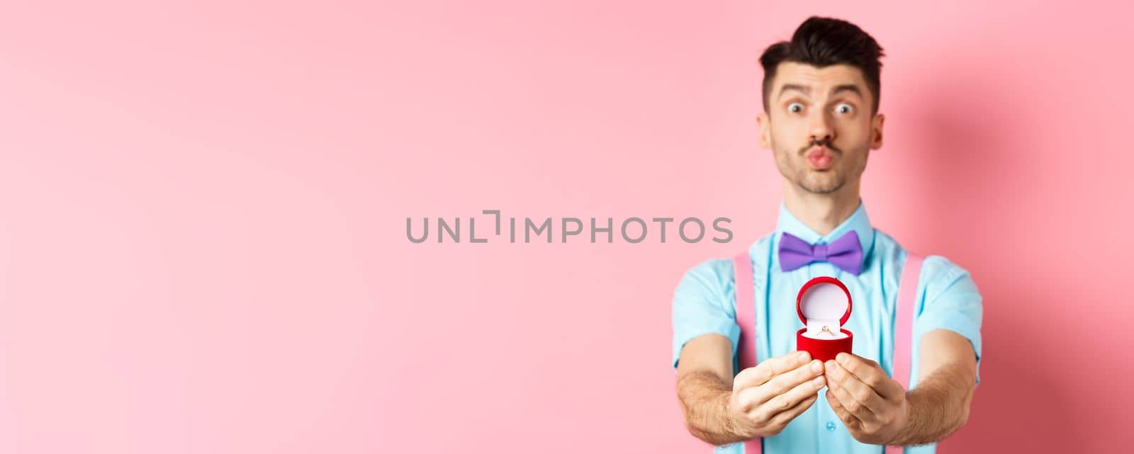 Valentines day. Funny young man pucker lips for kiss and showing engagement ring, making proposal, say marry me to lover, standing over pink background by Benzoix