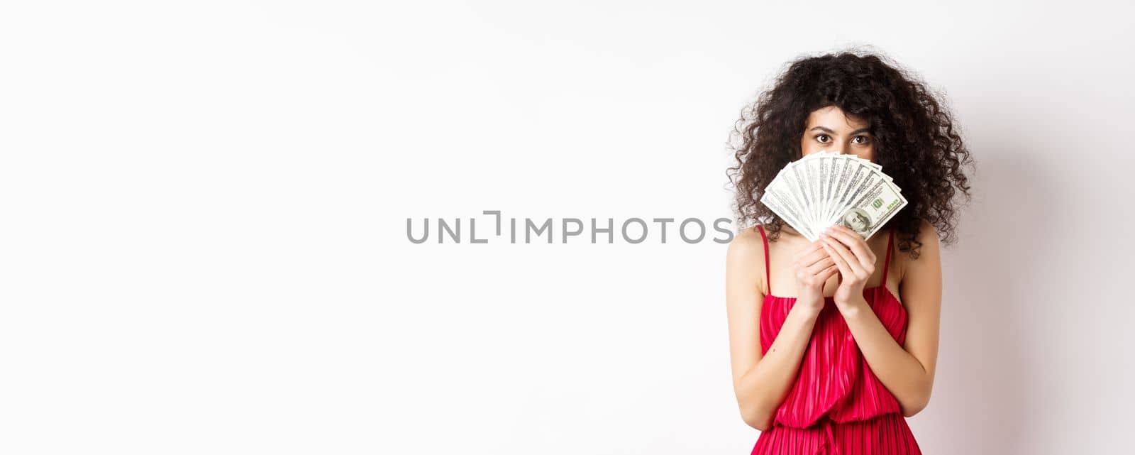 Gorgeous woman with curly hair, hiding face behind dollars and smiling, standing over white background. Copy space