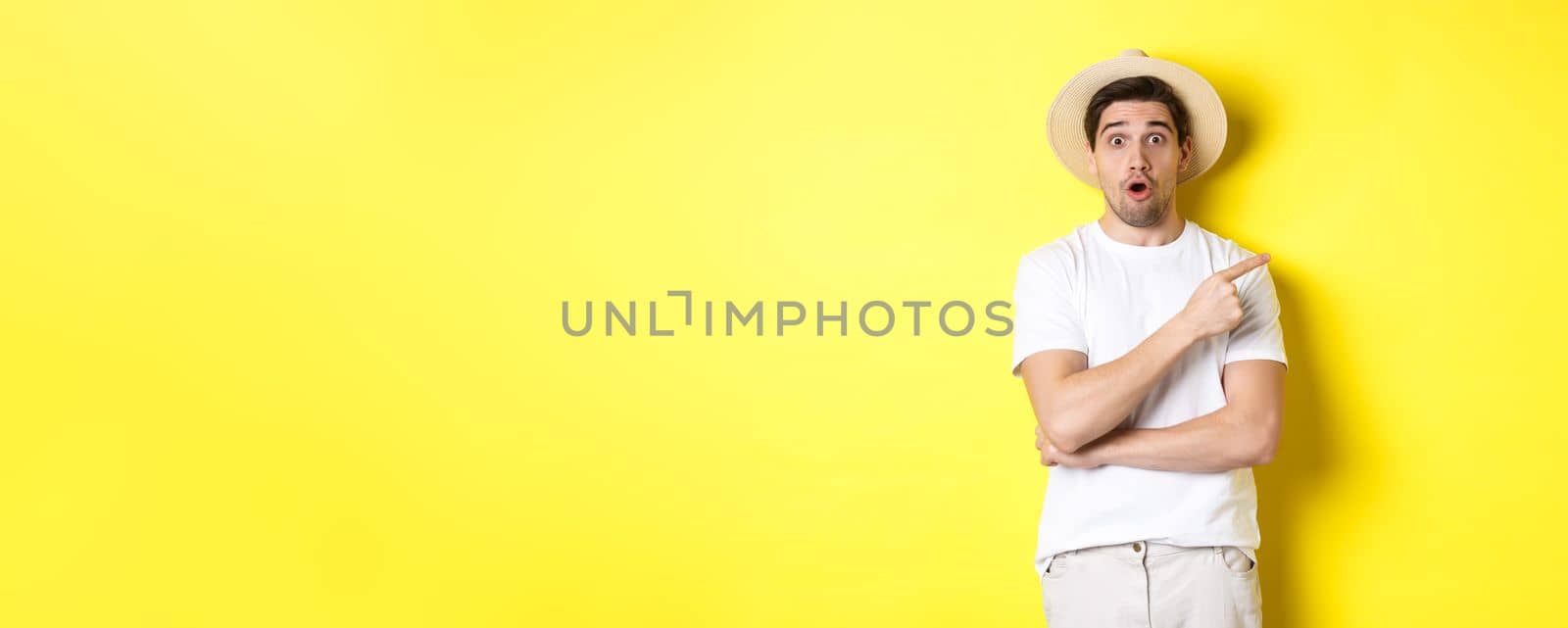 Surprised man in straw hat pointing finger right, showing promo banner, standing over yellow background. Copy space