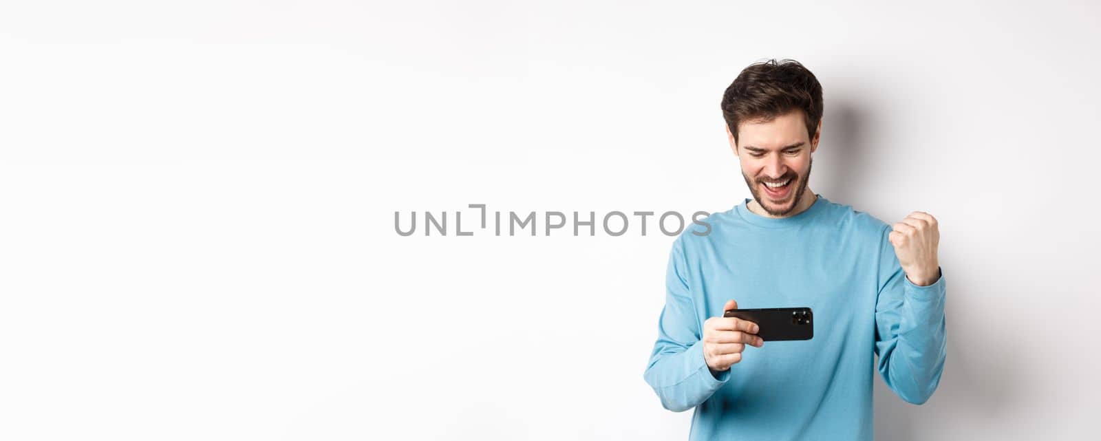 Happy young man winning in video game on smartphone, looking at mobile screen and say yes, making fist pump in celebration, achieve online goal, standing over white background.