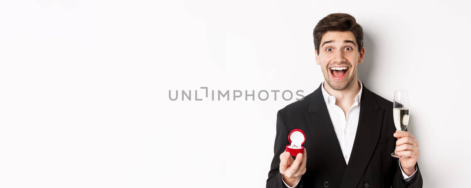Close-up of handsome man in suit, making a proposal, giving engagement ring and raising glass of champagne, standing against white background by Benzoix
