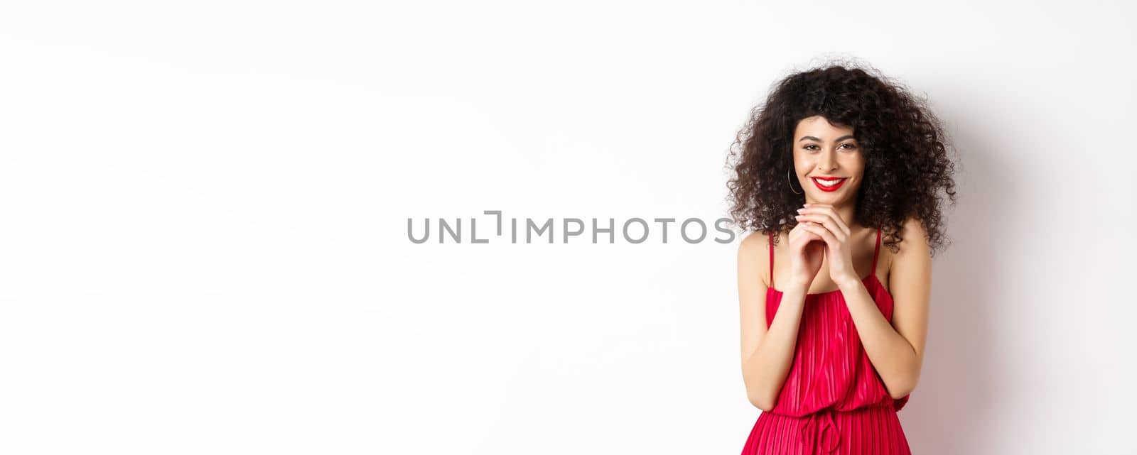 Cunning young woman with curly hair, steeple fingers and smiling devious, having a plan, standing in red dress on white background.