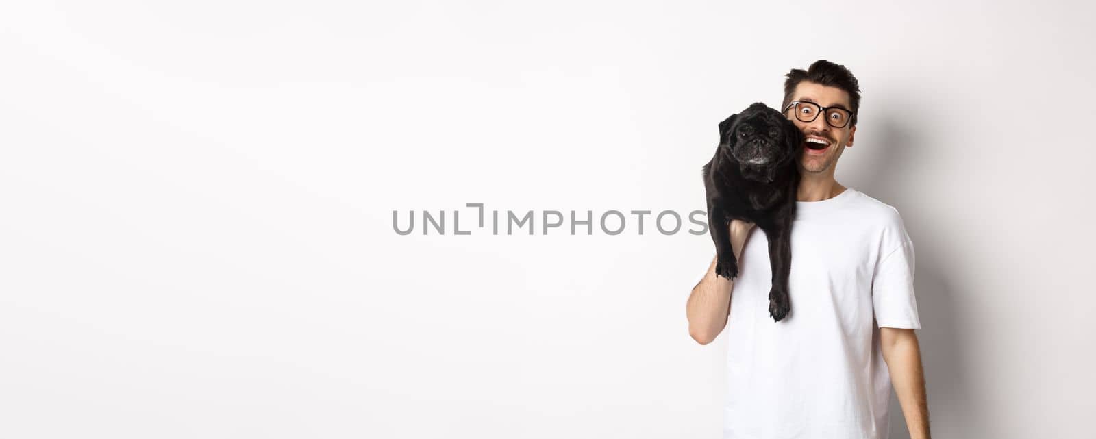 Cheerful hipster guy in glasses and t-shirt, carry cute black pug on shoulder and smiling. Dog owner playing with his pet, standing over white background.