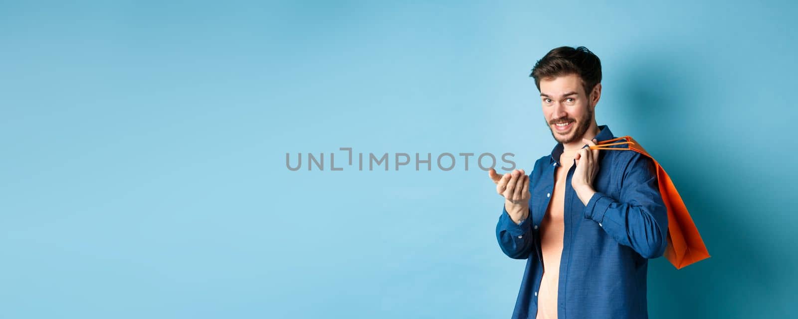 Modern guy holding shopping bag behind shoulder and beckon you, telling to come closer or inviting to store, standing on blue background by Benzoix