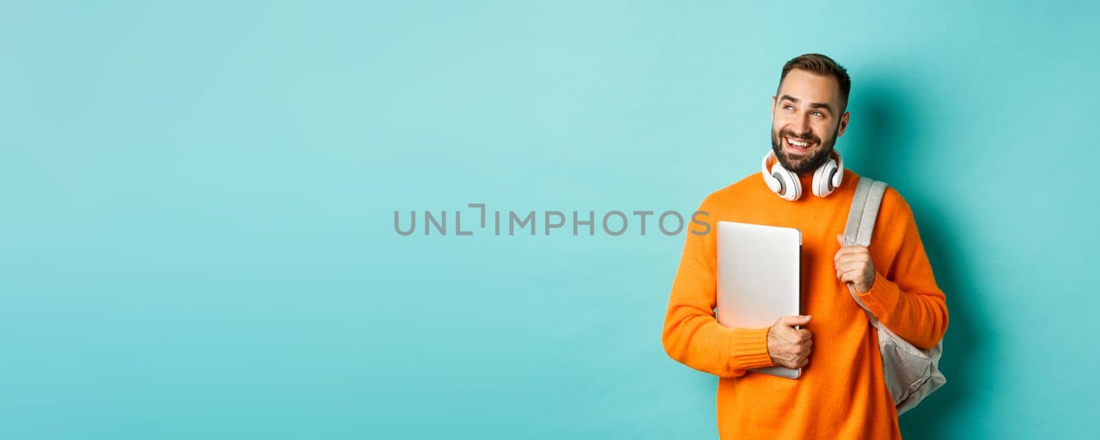 Happy man with backpack and headphones, holding laptop and smiling, looking left thoughtful, standing over turquoise background.