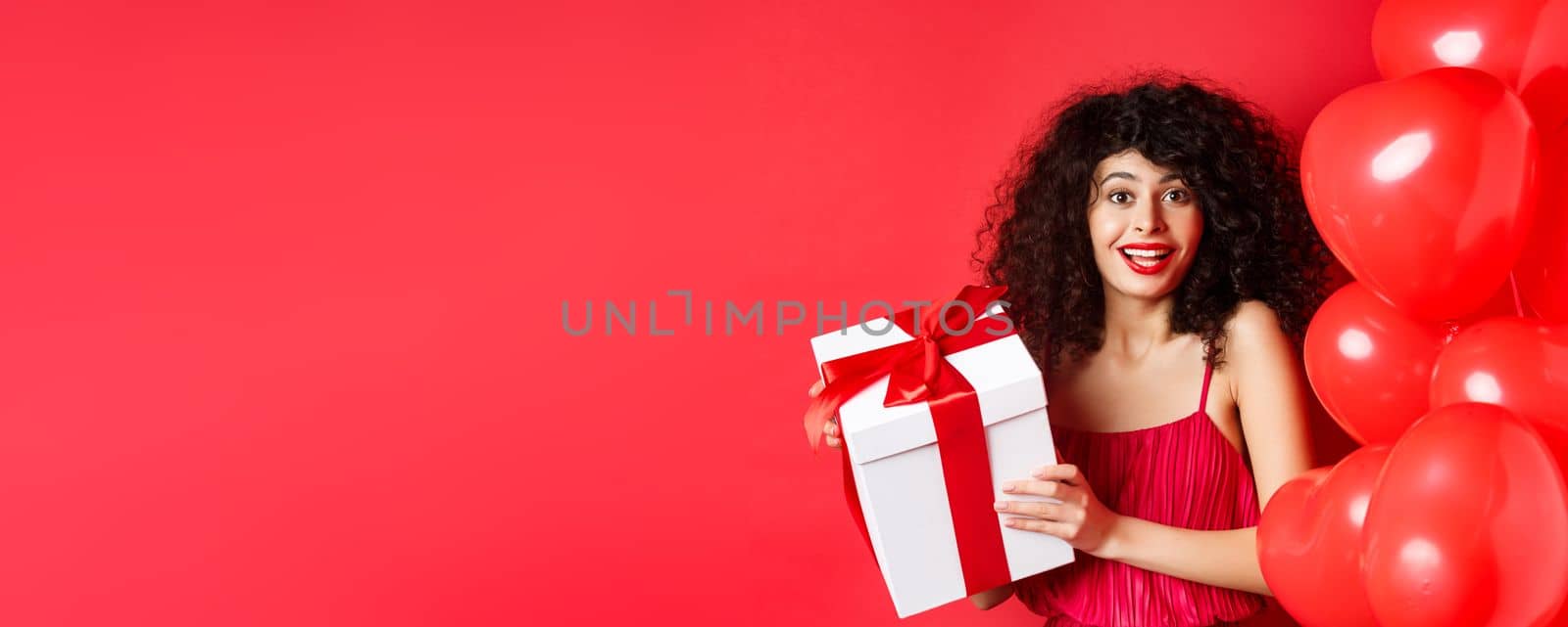 Holidays and celebration. Beautiful woman with curly hair, standing near heart balloons, holding gift box and smiling happy, white background.