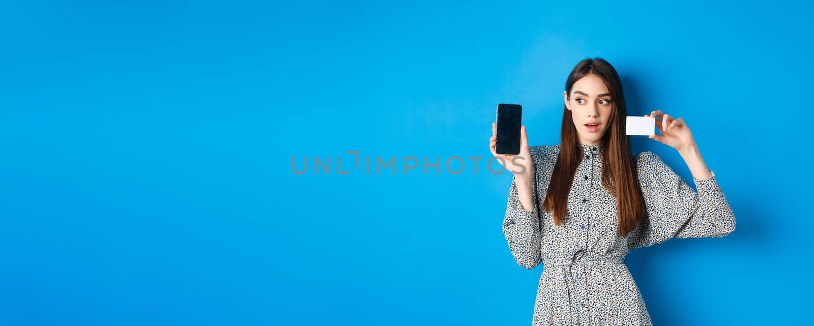 Online shopping. Pensive lady in dress showing empty phone screen and plastic credit card, order in internet, blue background.