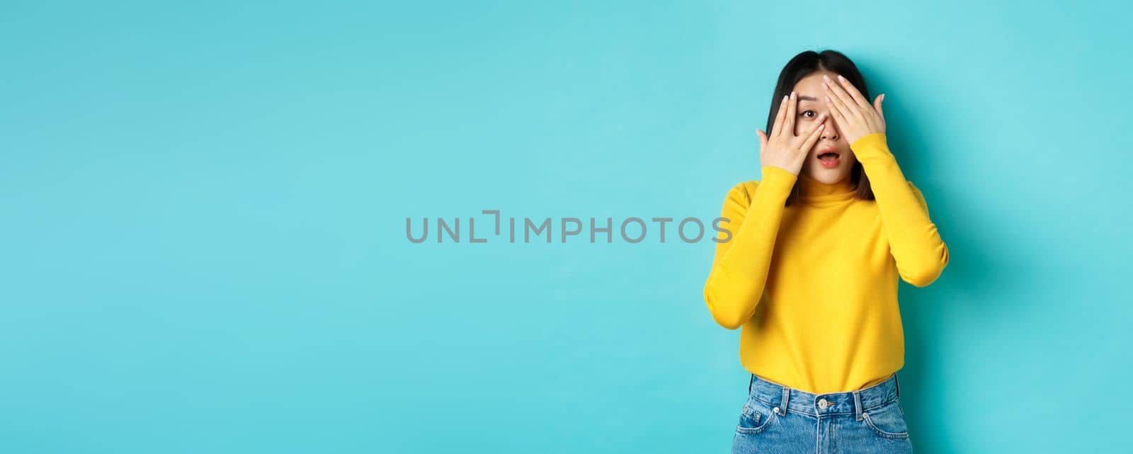 Intrigued asian girl peeking through fingers at something interesting, looking curious at camera, standing over blue background by Benzoix