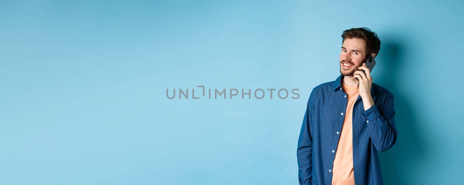 Modern smiling guy talking on mobile phone, looking happy at camera, standing on blue background.