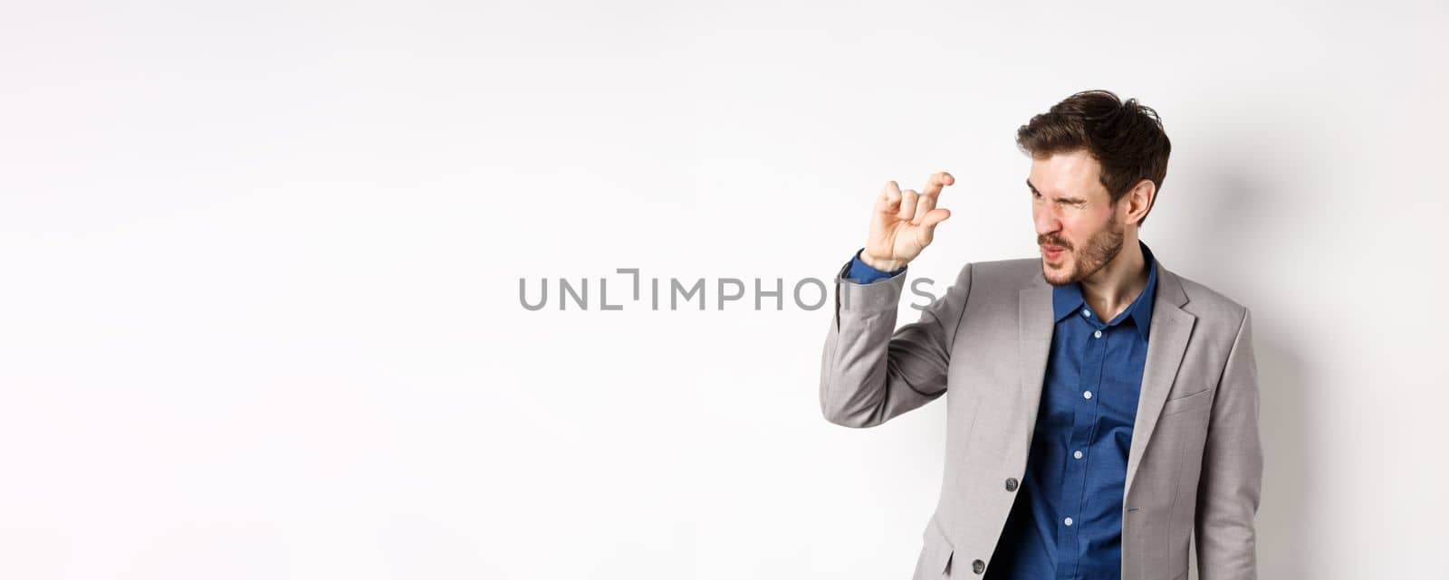 Businessman trying to see something tiny, showing little thing size with fingers, standing in suit on white background by Benzoix