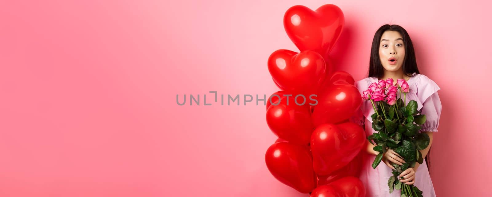 Surprised asian girl in dress standing near valentines day heart balloons and say wow at camera, holding flowers bouquet from lover, romantic date with roses, pink background.