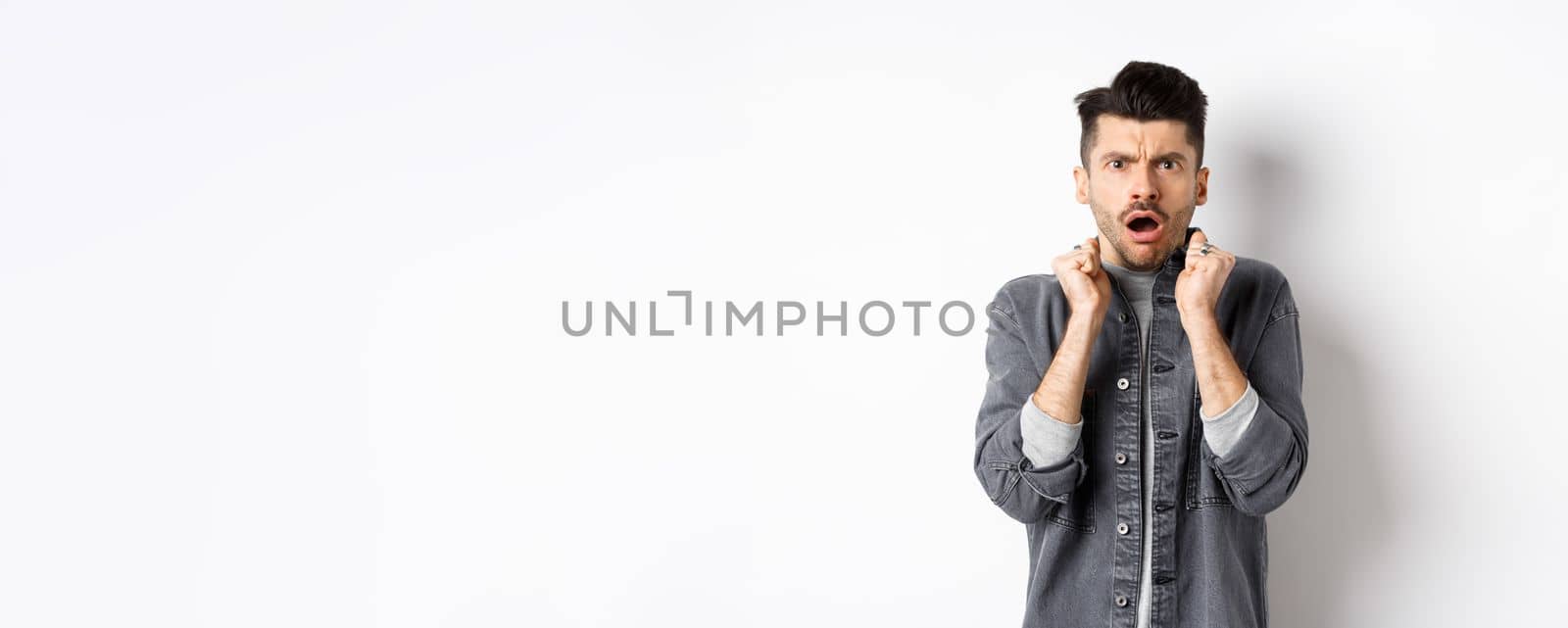 Shocked young man look confused and offended, gasping and frowning at camera, hear bad news, standing on white background.