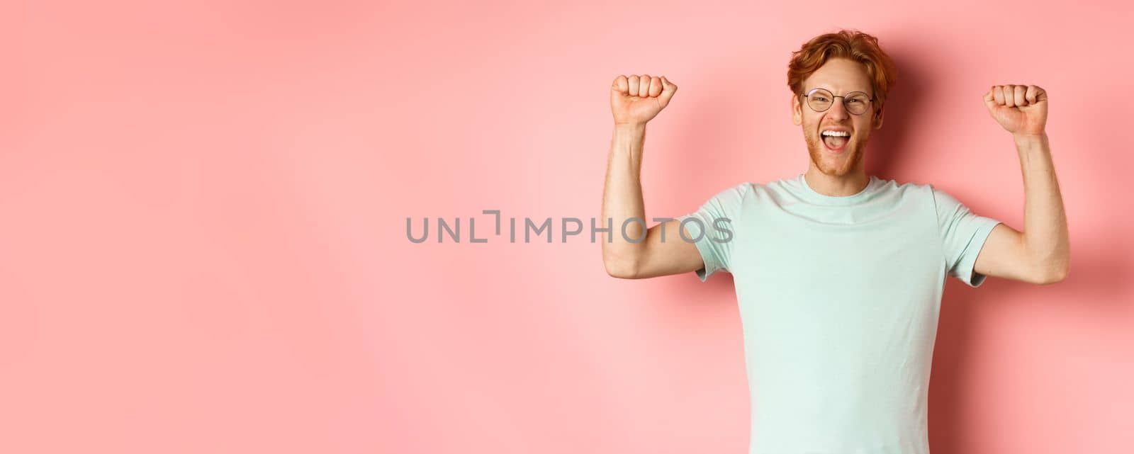 Happy winner with red hair and beard celebrating victory, shouting yes with joy and raising hands up, enjoy success, standing over pink background by Benzoix