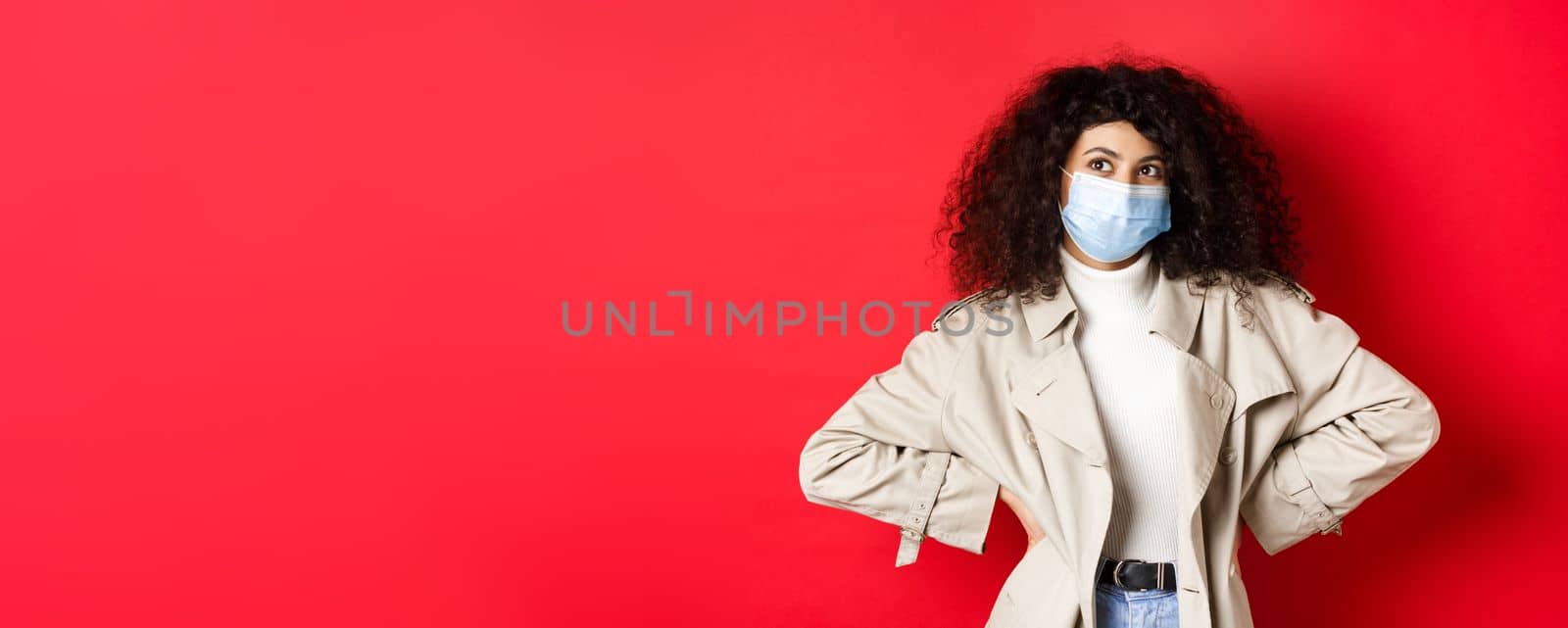 Covid-19, pandemic and quarantine concept. Stylish enthusiastic woman in medical mask and trench coat, smiling and looking at upper left corner, red background by Benzoix