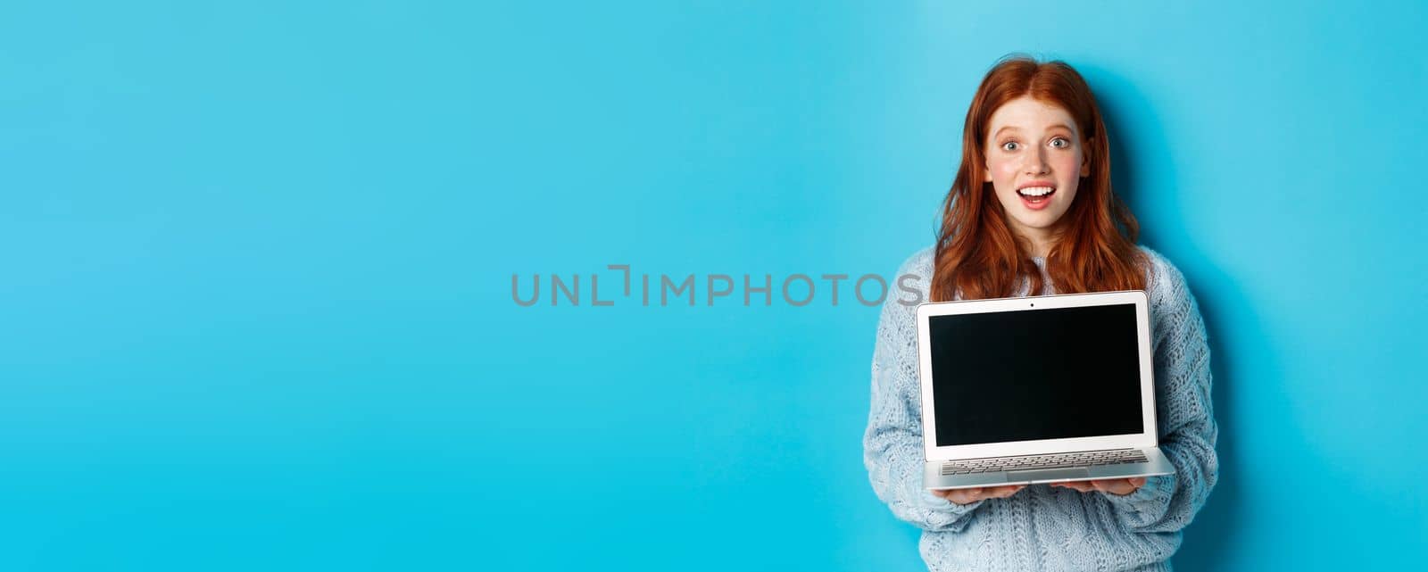 Excited redhead female freelancer showing laptop screen, staring at camera amazed, standing with computer against blue background.