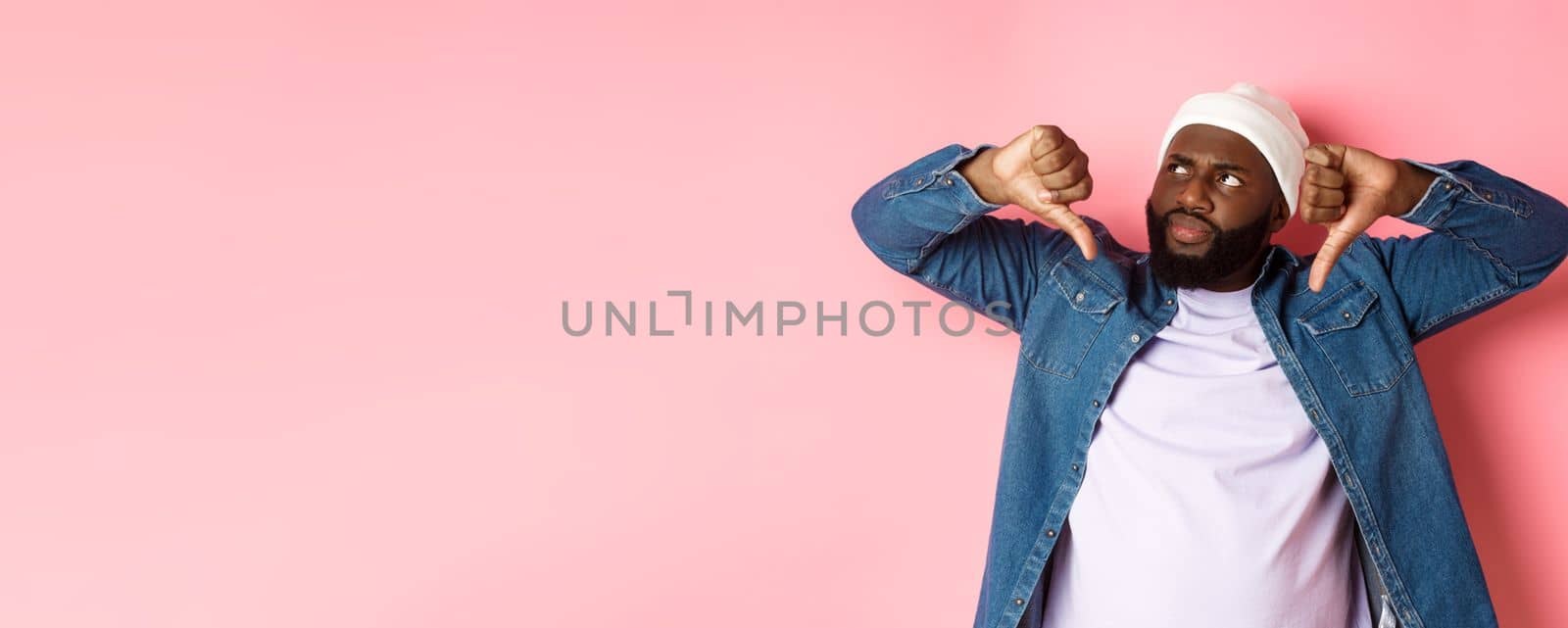 Disappointed Black man staring at upper left corner annoyed, showing thumbs-down, dislike something, standing over pink background by Benzoix