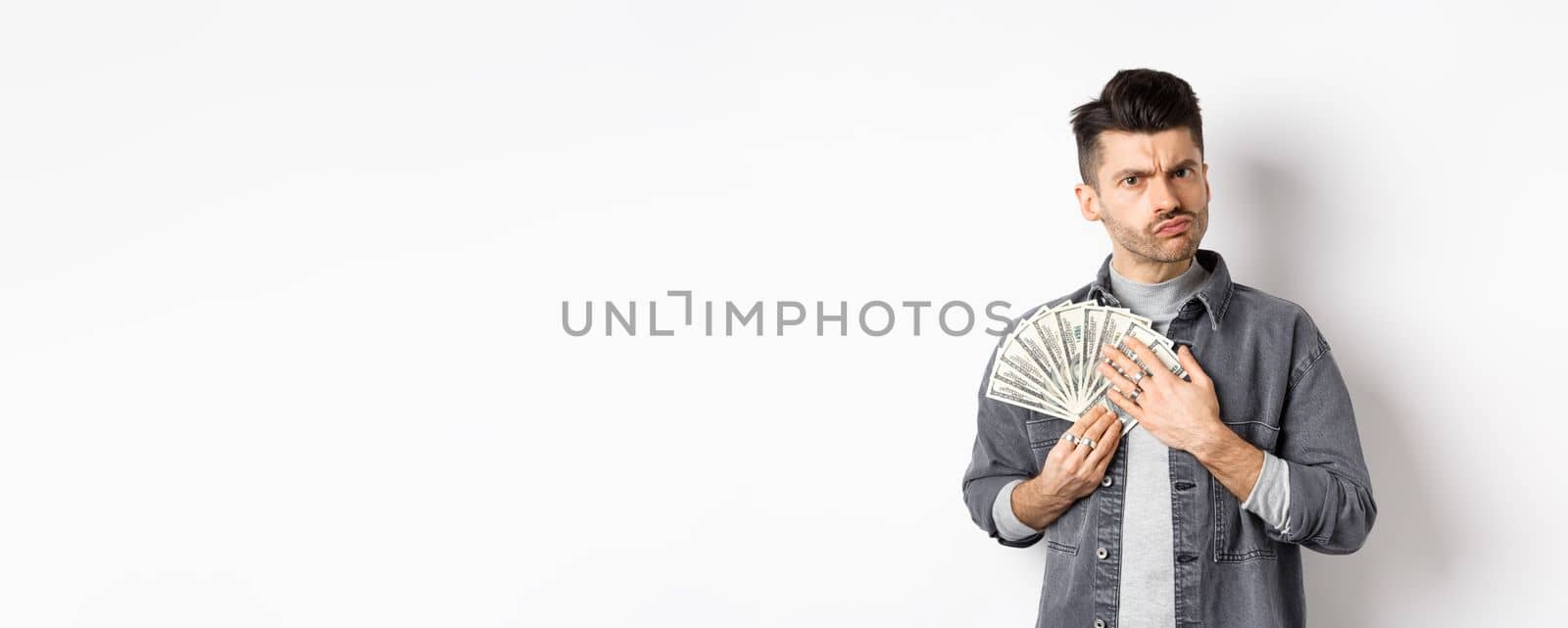 Greedy guy frowning and hugging dollar bills, unwilling to share money, standing on white background.