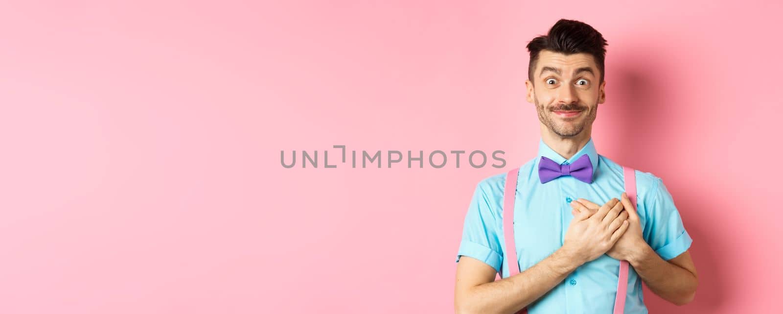 Cheerful young man with moustache, wearing shirt and bow-tie, holding hands on heart and smiling grateful, saying thank you, standing on pink background.