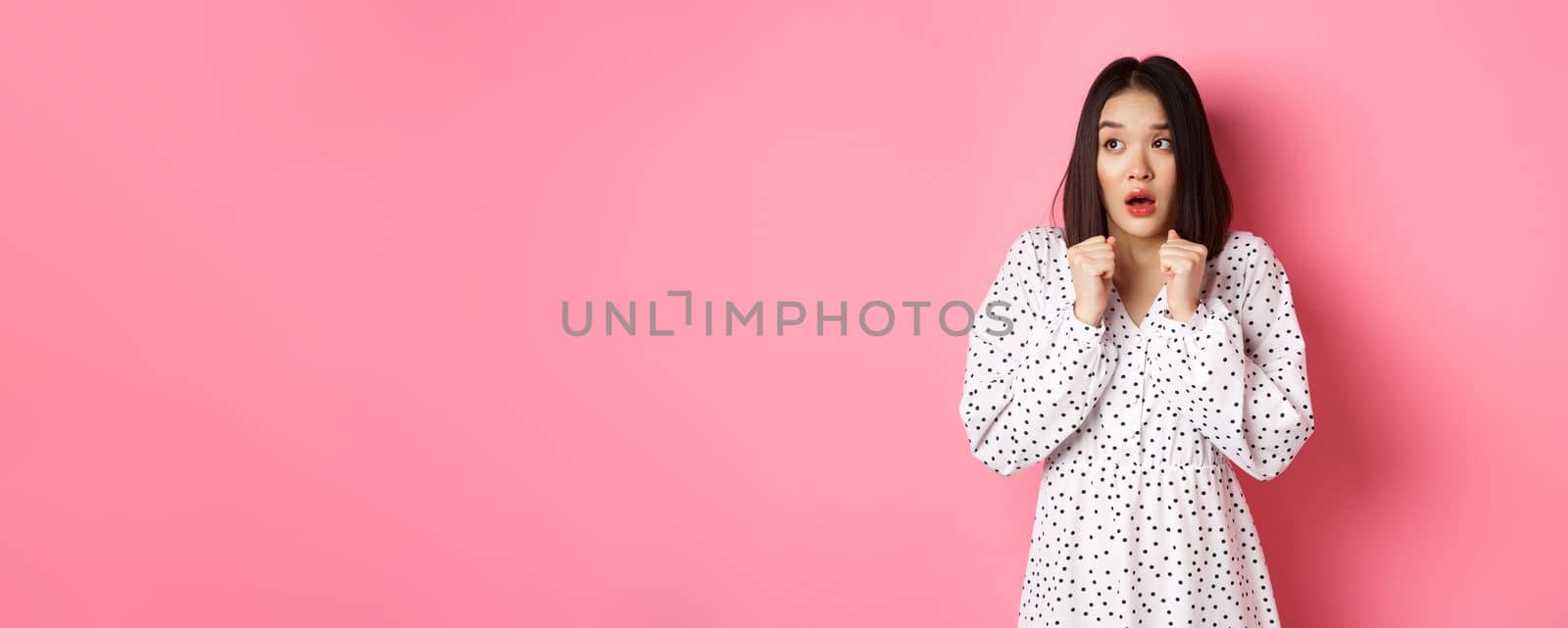 Scared and timid asian girl trembling from fear, staring left and gasping, standing in dress over pink background.
