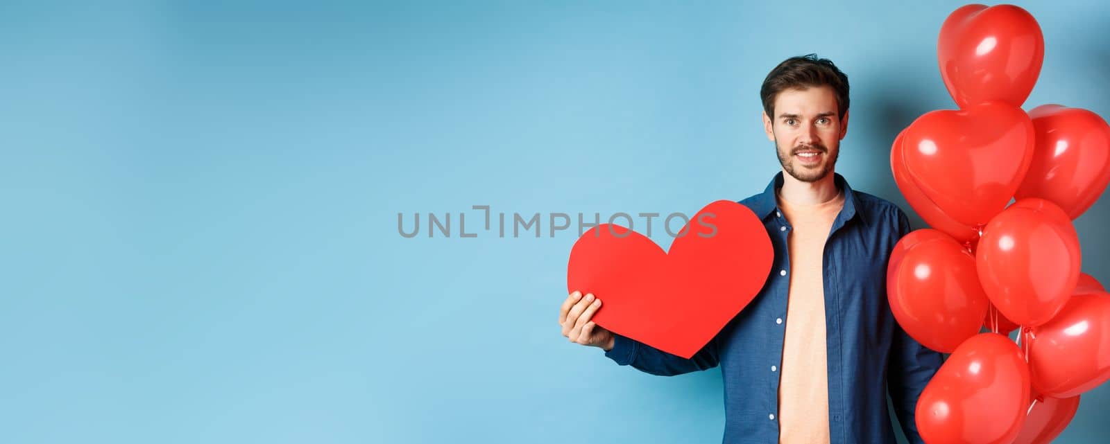 Valentines day and love concept. Smiling young man waiting for lover with romantic gifts, heart balloons and valentine postcard, standing over blue background.
