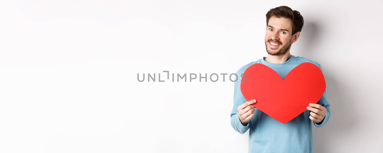 Handsome european man in sweater saying I love you, boyfriend standing with valentines day red heart, posing over white background by Benzoix