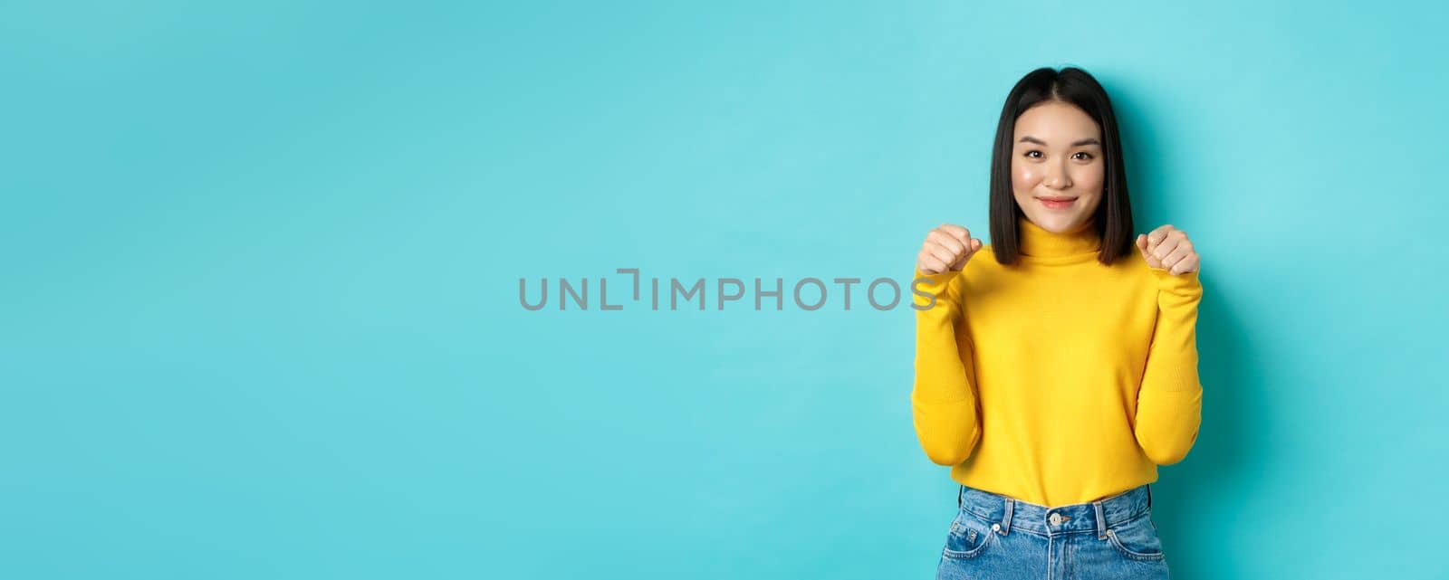 Beauty and fashion concept. Beautiful and stylish asian woman in yellow pullover, holding hands raised near chest as if holding banner or logo, standing over blue background.