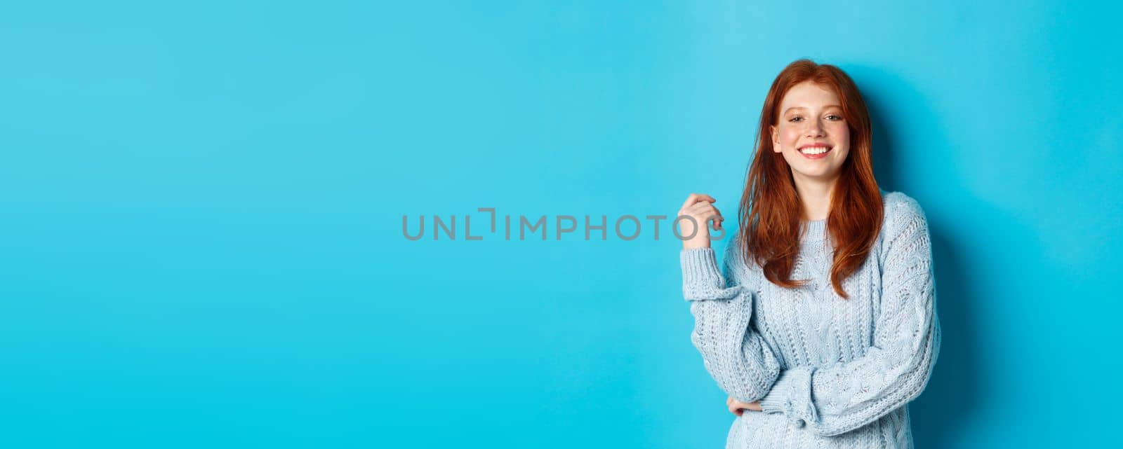 Happy redhead female in sweater, looking pleased at camera and smiling, standing against blue background.