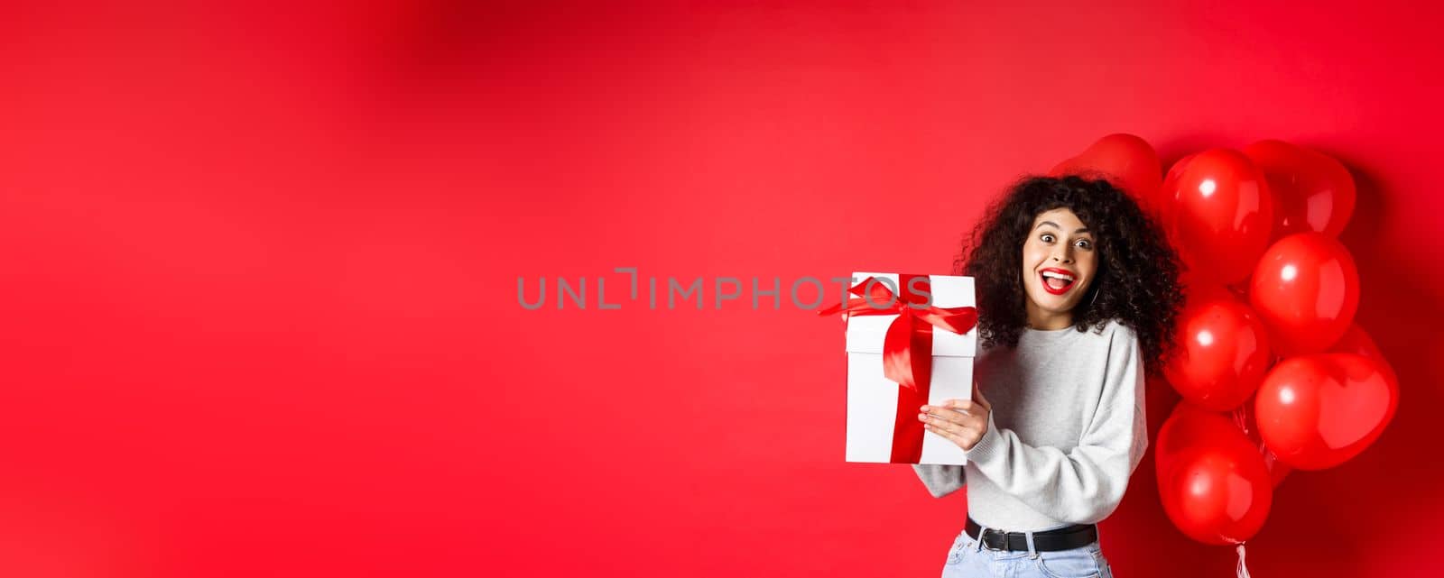 Surprised and happy woman holding valentines day gift from lover, standing near romantic hearts balloons and looking at camera amazed, red background.