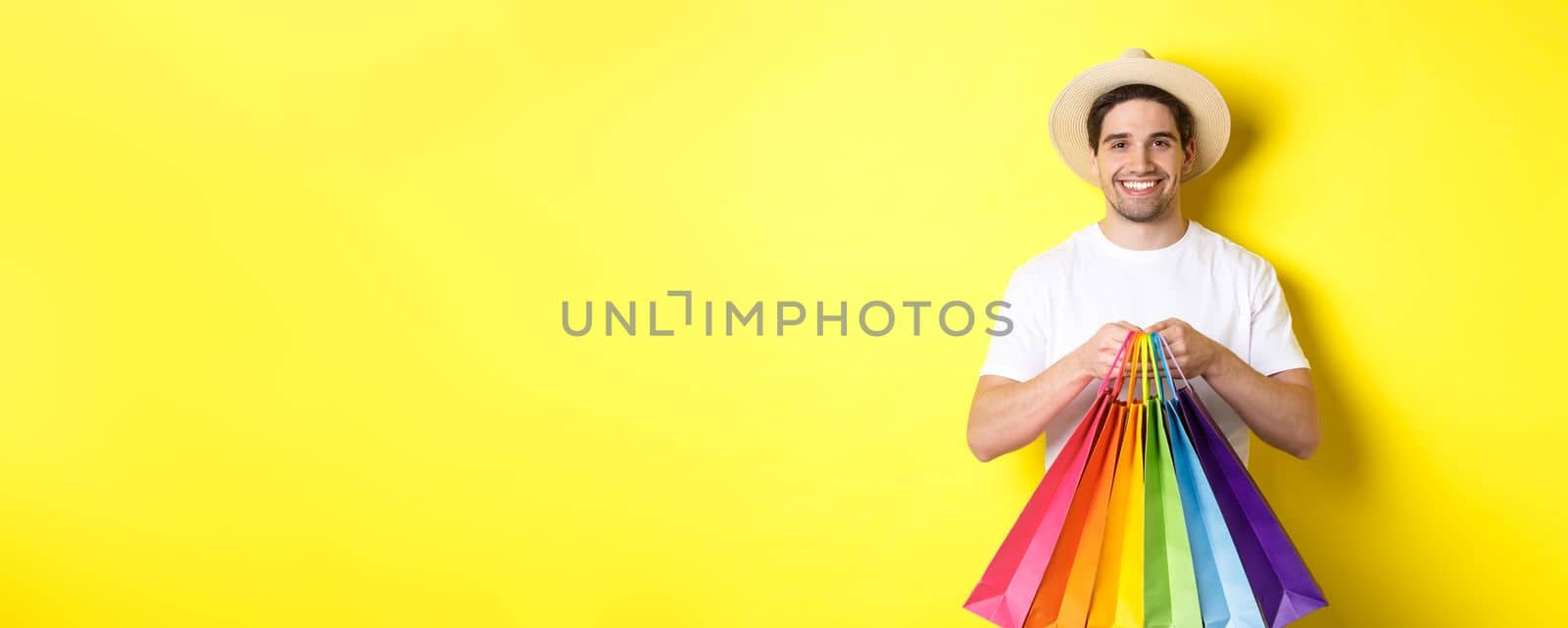 Image of happy man shopping on vacation, holding paper bags and smiling, standing against yellow background.