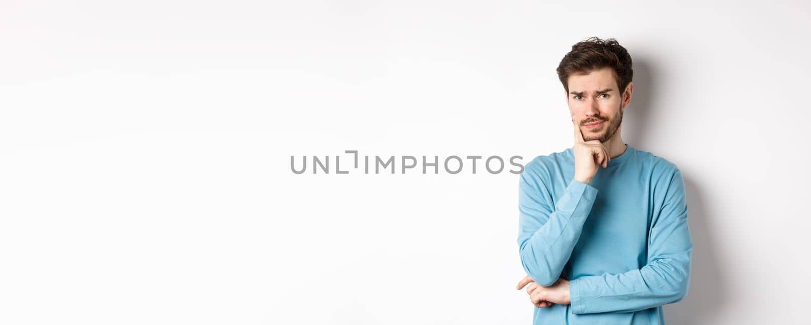 Skeptical young man frowning, looking with worried thoughtful look, standing upset over white background.